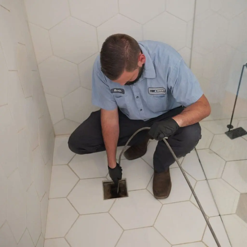 Floor drain cleaning process in Phoenix, Arizona with a drain auger cable inserted into a tiled shower drain to remove blockages. Day & Night Air Conditioning, Heating, and Plumbing provides commercial drain cleaning in Phoenix, helping businesses clear clogged drains and maintain proper drainage systems. The setup shows real drain service work using a flexible cable to break up debris and restore water flow. Businesses in Phoenix can contact Day & Night Air Conditioning, Heating, and Plumbing at (602) 584-7758 for commercial drain cleaning services, including maintenance and blockage removal.