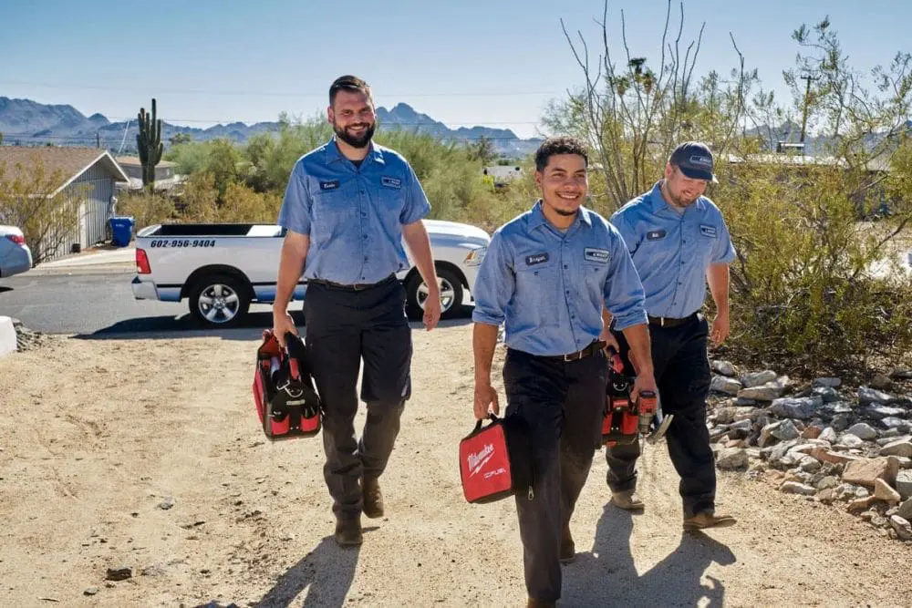 Day & Night Air Conditioning, Heating, and Plumbing technicians walking through a Scottsdale, Arizona neighborhood while carrying HVAC tools and equipment. The scene represents HVAC services in Scottsdale, where trained technicians travel to properties to handle air conditioning repair, heating system installation, and routine maintenance. The desert surroundings, service truck, and equipment highlight real service conditions in the area. Customers can contact Day & Night Air Conditioning, Heating, and Plumbing at (602) 584-7758 for HVAC services in Scottsdale, including repair, installation, and maintenance.